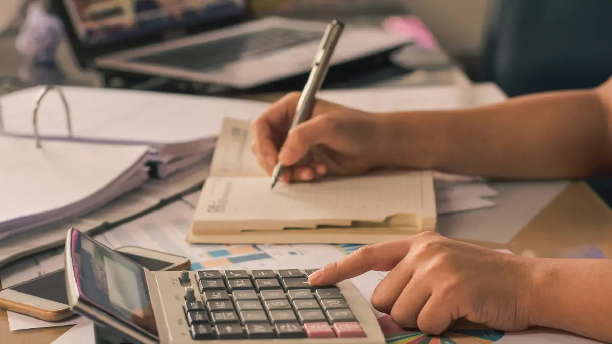 Hands writing in a notebook beside a calculator and laptop while comparing costs