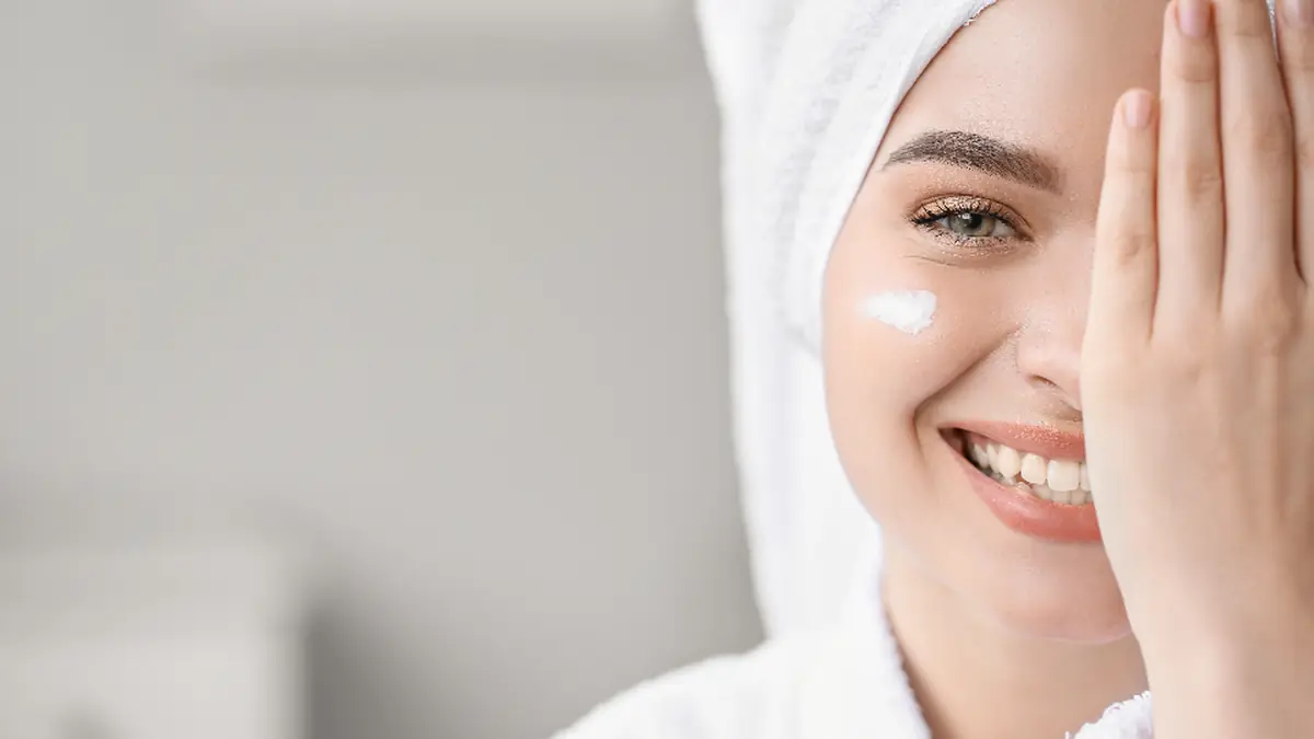 Woman with a towel on her head applying skincare product to her face